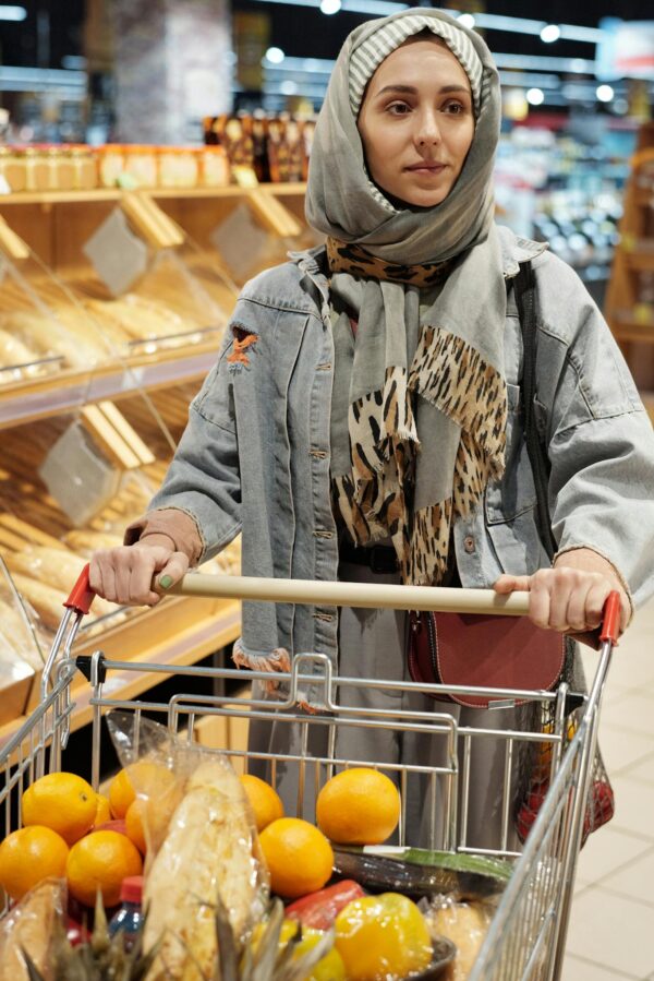 Woman in hijab shopping at a supermarket with a cart full of groceries and fruits.
